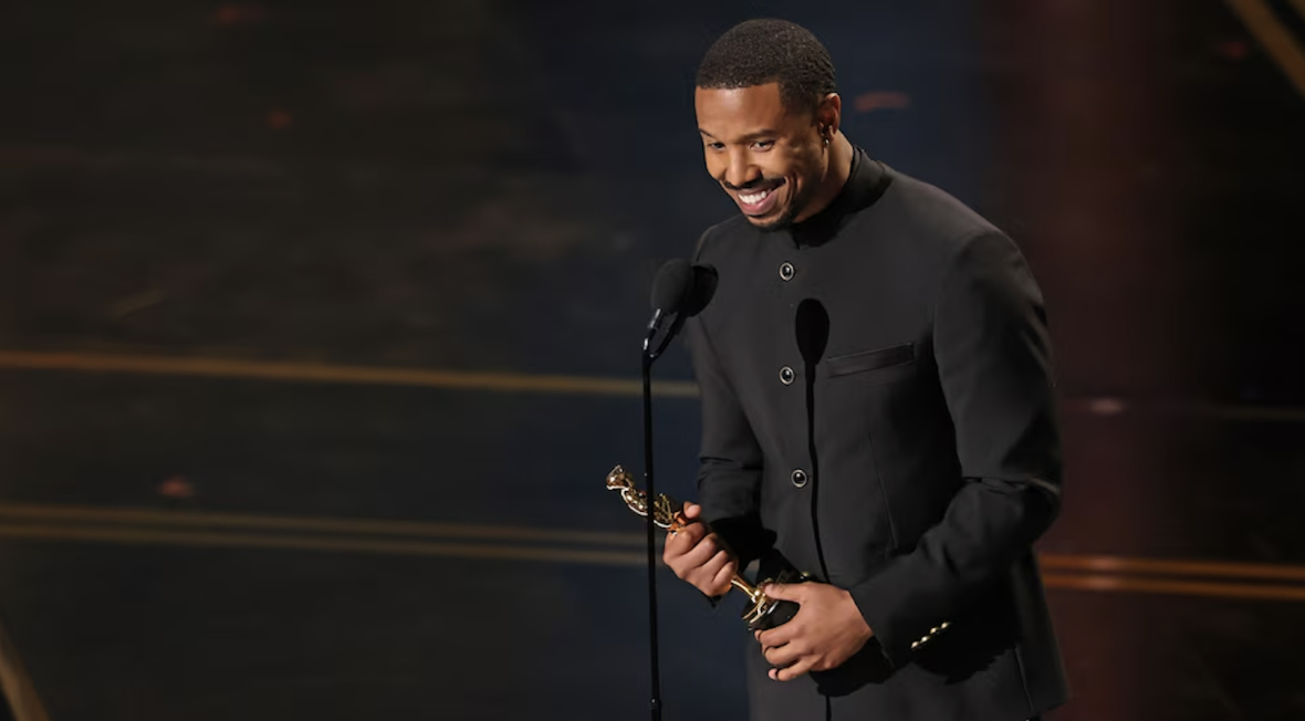 Michael B. Jordan smiles at the microphone while holding his Oscar onstage, dressed in a black high-collar suit against a dark, softly lit background.