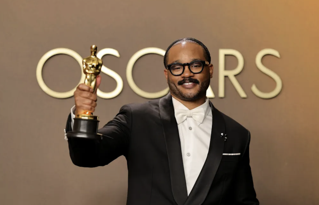 Ryan Coogler, wearing a black tuxedo and white bow tie, holds up an Oscar statuette while posing in front of an Oscars backdrop.